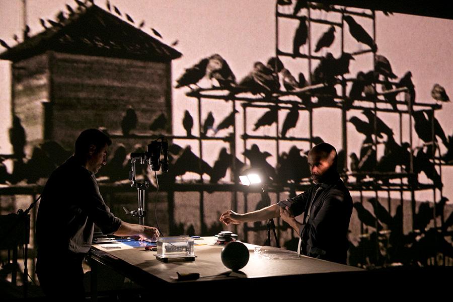 Photo from stage production of 'Birdie' featuring a projection of Hitchcock's 'The Birds' in the background while artists work with small objects on a table