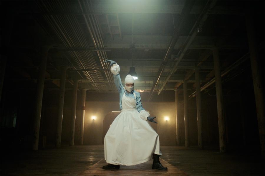 Film still from Black Lodge featuring image of a performer in white striking a pose in a dimly lit room