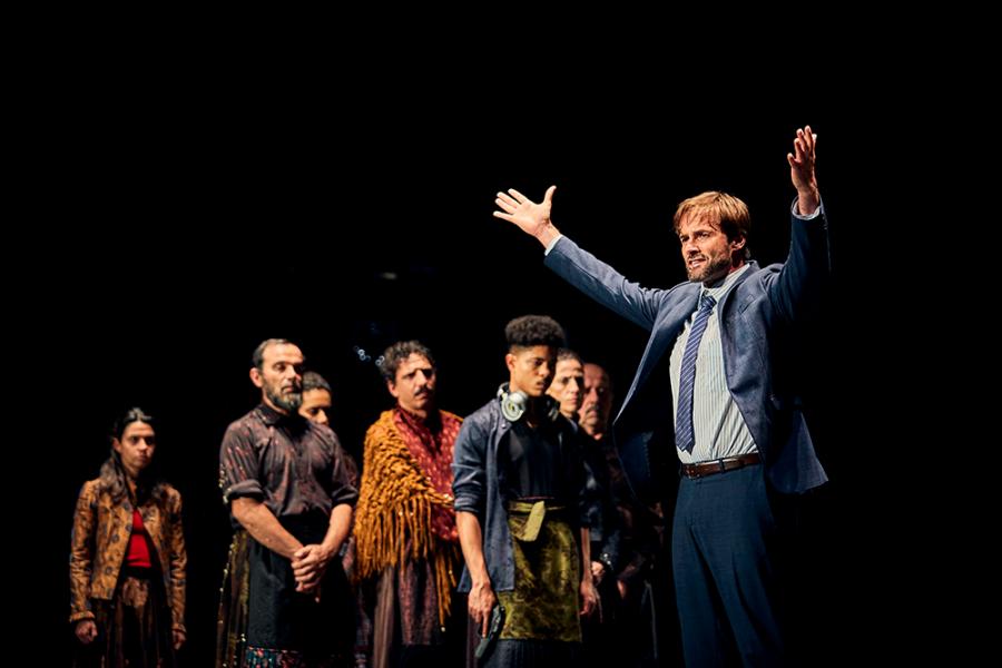 A performer on stage poses with outstretched arms and cast members in the foreground. 