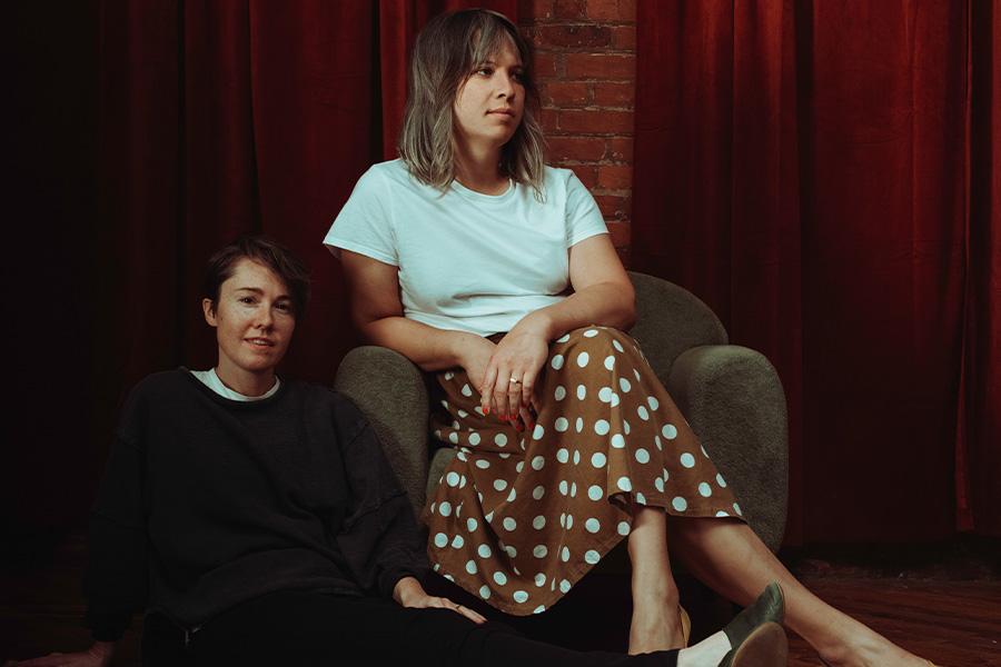 Andrew Yee and Caroline Shaw pose together in front of 2 red velvet curtains and a strip of brick wall. Shaw sits on the floor to the left of Yee, smiling slight at the camera and wearing a tee shirt layered underneath a grey sweater and black pants. Yee sits in a chair next to Shaw, wearing a white tee and a brown skirt with white polka dots.