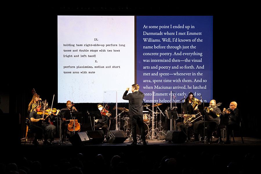 An orchestra performs on stage in front of a screen projecting text
