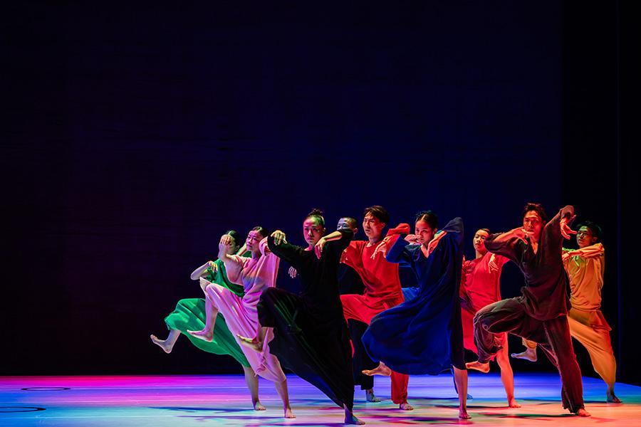 Members of Cloud Gate march across a rainbow lit stage wearing colorful gowns. They move in unison with one leg raised, mid large step.