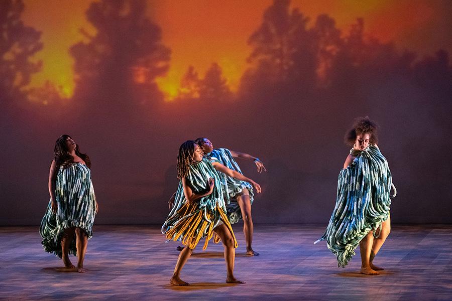 four dancers on stage in cultural dress with a fiery backdrop with trees