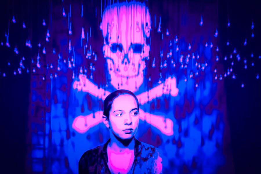 A woman stands in front a projection of a skull and bones, cast onto a sheet, illuminated by purple and blue light.