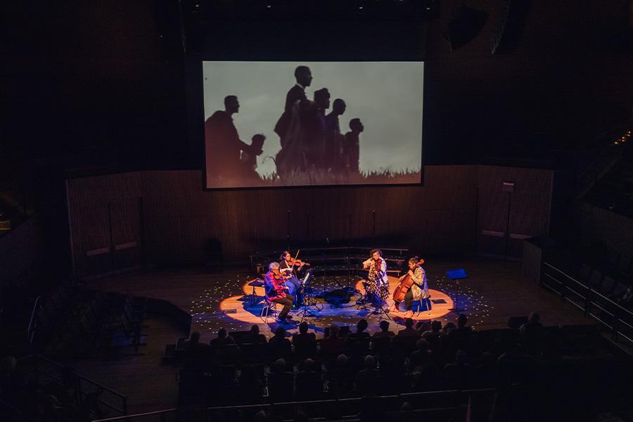 birds eye view of Kronos quartet members performing on stage amongst an audience with a projection in the background