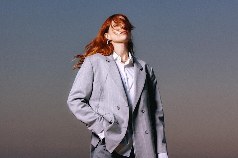 Stylized portrait of Laurel Halo in a light gray oversized suit, standing confidently against a dusk sky with windblown hair partially obscuring her face — evoking a mood of elegance, individuality, and artistic cool