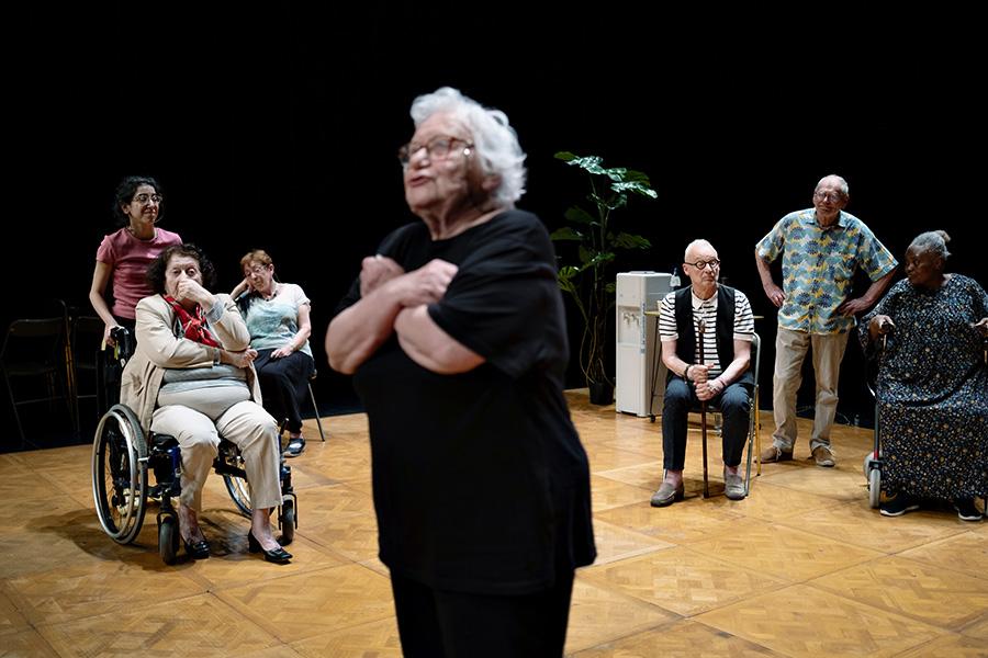 The cast of La Vie Secrète des Vieux performs on stage. An elderly woman in all black and wearing glasses stands in the center, speaking and looking off to the left. She is slightly blurred as the camera focuses on her fellow performers in the background, watching as she performs, 5 people seated and 2 standing.