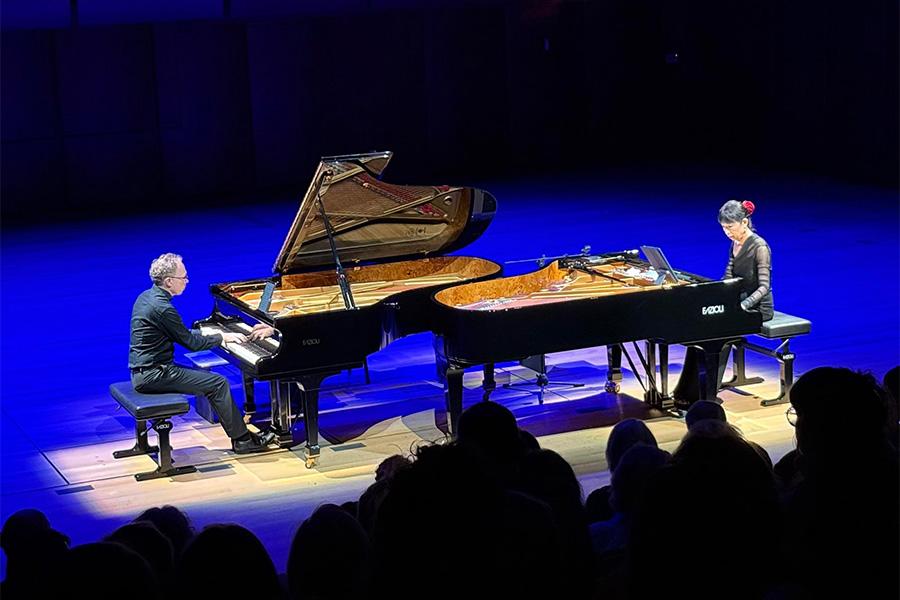 Gloria Cheng and Ralph van Raat perform on stage, lit by blue lighting. Both artists perform at their own pianos, positioned to be facing one another. The photo is taken from a high angle, showing the silhouettes of some audience members in the foreground.