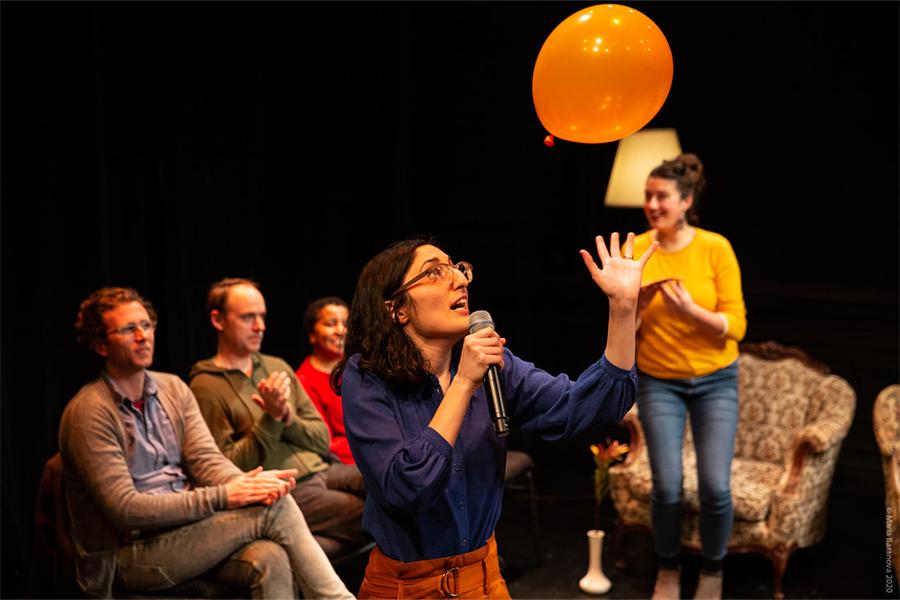 Image of performer on stage with audience members seated in chairs also on stage, watching as the performer looks at an orange balloon