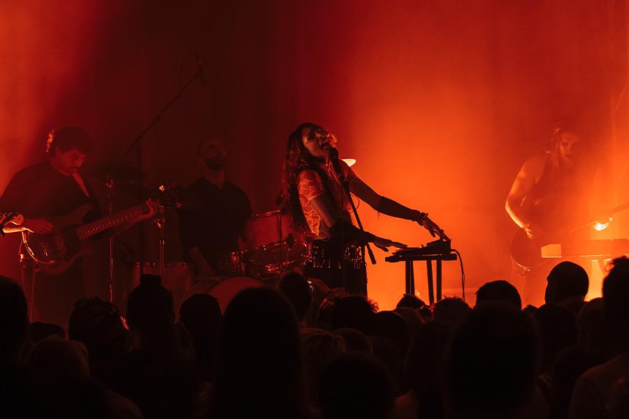 SPELLLING performs on stage, lit by orange light. There are the silhouettes of audience members in the foreground and blurred silhouettes of SPELLLING's band behind them performing on stage.