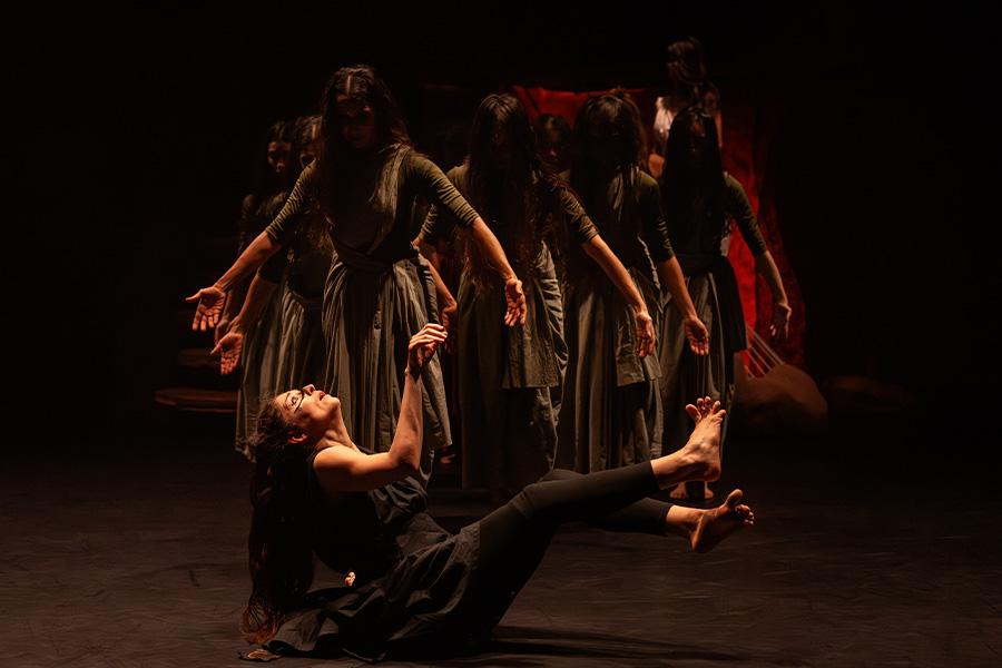 A group of dancers stand with arms partially stretched out to their sides on a dark stage wearing grey dresses. They look downwards at another dancer, wearing all black, seated on the floor with her arms and legs extended up and outward, looking startled.