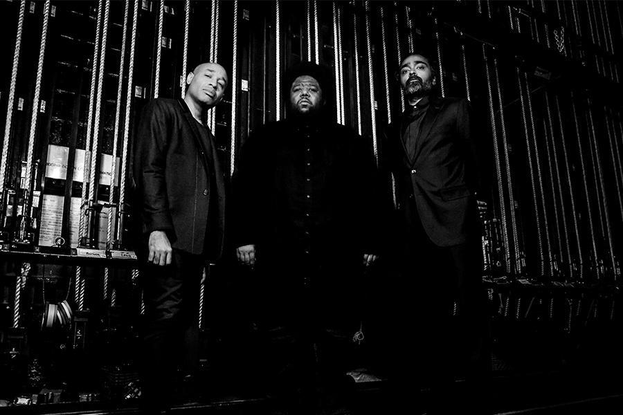 Black and white portrait of band members dressed in dark suits standing in a backstage theater setting, surrounded by stage rigging and ropes