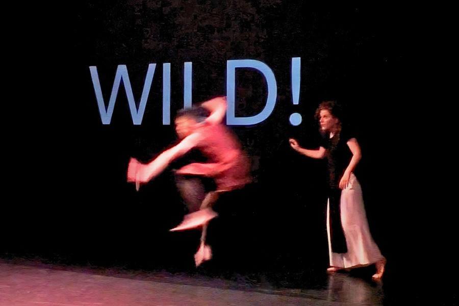 Two dancers perform in front of black back drop with the world 'WILD!' projected onto it. One of the dancers wears a red shirt and jumps mid air while the other performer steps towards them with one arm outward.