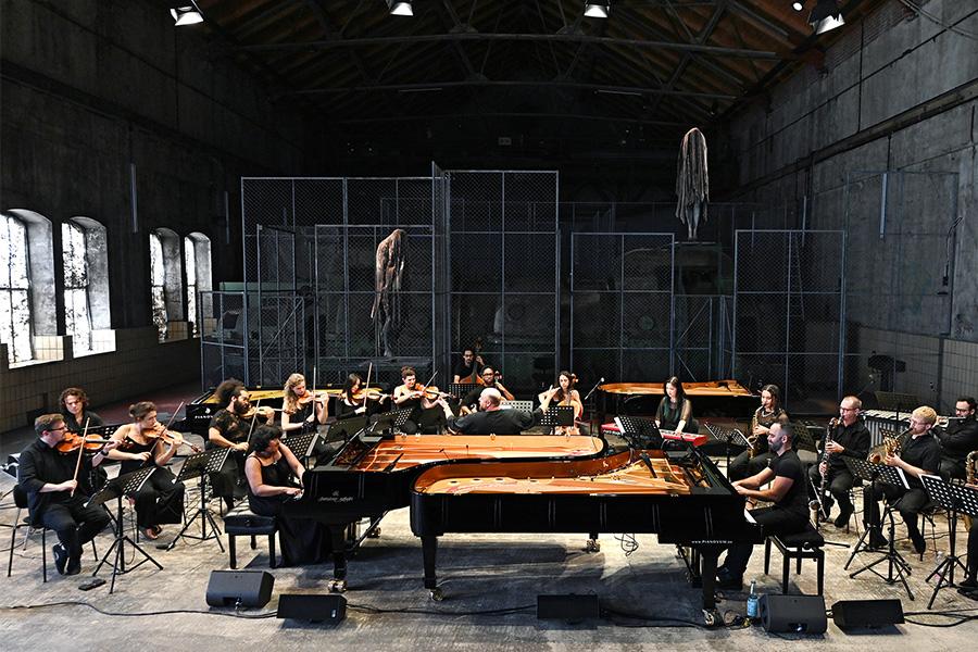 Musicians perform at two pianos, surrounded by a large orchestra, in a warehouse like space