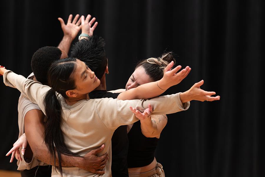 Four contemporary dancers embrace during a rehearsal, creating a sculptural moment of movement, connection and collaboration in a performing arts studio.