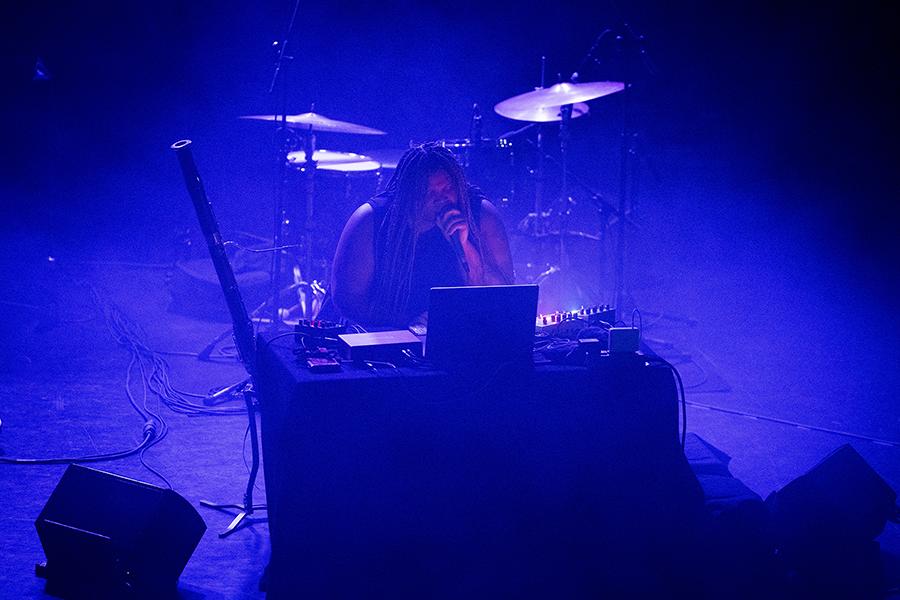 Under moody blue lighting, Joy Guidry commands the stage behind a table filled with electronic gear, passionately performing with a microphone in hand, surrounded by cables, a laptop