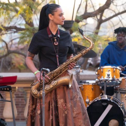 A performer stands on stage, looking to their right and holding a saxophone