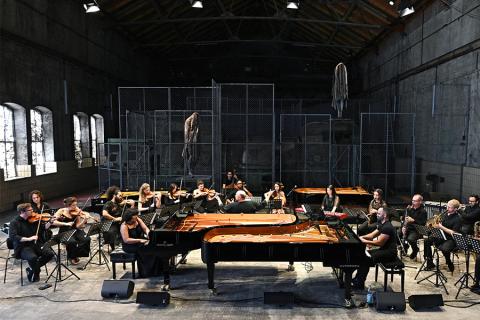 Musicians perform at two pianos, surrounded by a large orchestra, in a warehouse like space
