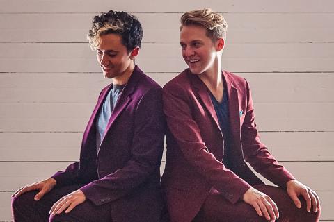 Caleb Teicher and Nic Gareiss pose together in maroon suits, seated next to each other with their backs to one another. Teicher looks towards the ground while smiling and Gareiss looks over their shoulder towards Teicher smiling.