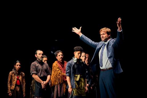 A performer on stage poses with outstretched arms and cast members in the foreground. 