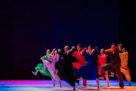 Members of Cloud Gate march across a rainbow lit stage wearing colorful gowns. They move in unison with one leg raised, mid large step.