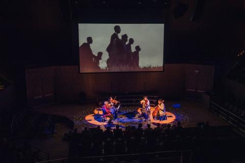 birds eye view of Kronos quartet members performing on stage amongst an audience with a projection in the background