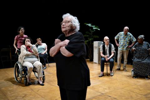 The cast of La Vie Secrète des Vieux performs on stage. An elderly woman in all black and wearing glasses stands in the center, speaking and looking off to the left. She is slightly blurred as the camera focuses on her fellow performers in the background, watching as she performs, 5 people seated and 2 standing.