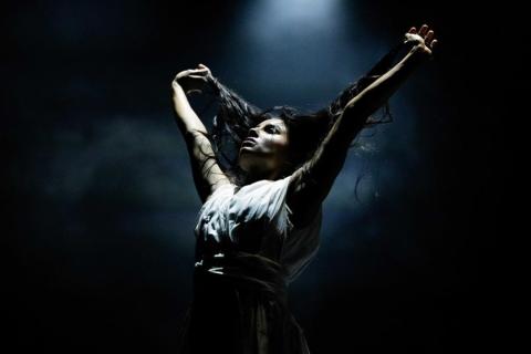 A dancer poses with arms raised above their head on a stage with a dark backdrop and smoke floating in the air.