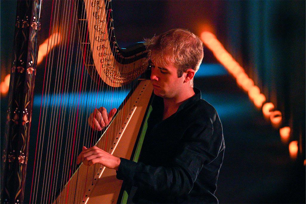 Parker Ramsay wearing a black shirt playing the harp on stage with blue lighting