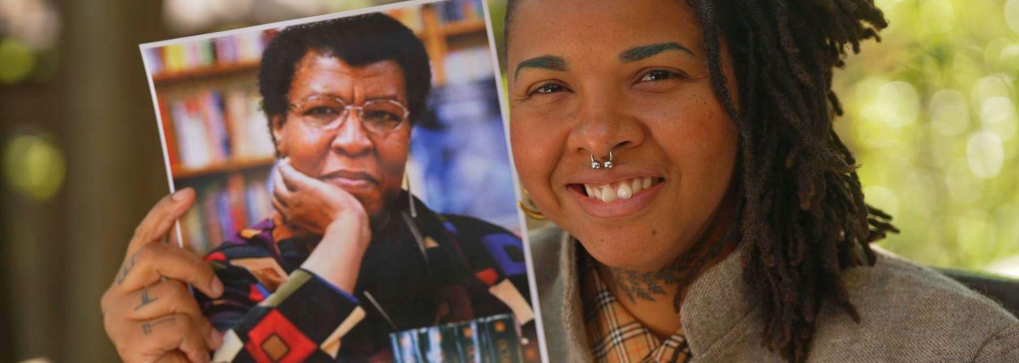 Madin Lopez smiles at the camera and holds up a photo of Octavia Butler. Trees are in the background.