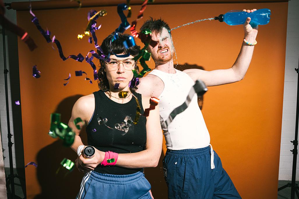 Two performers from Burnout Paradise pose in front of an orange backdrop while confetti falls around them