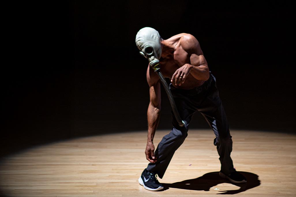 David Rousseve strikes a pose on stage in front of a black backdrop while wearing a gas mask 