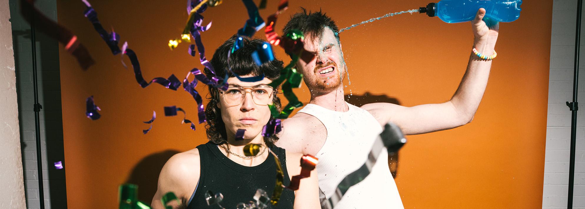 Two performers from Burnout Paradise pose in front of an orange backdrop while confetti falls around them