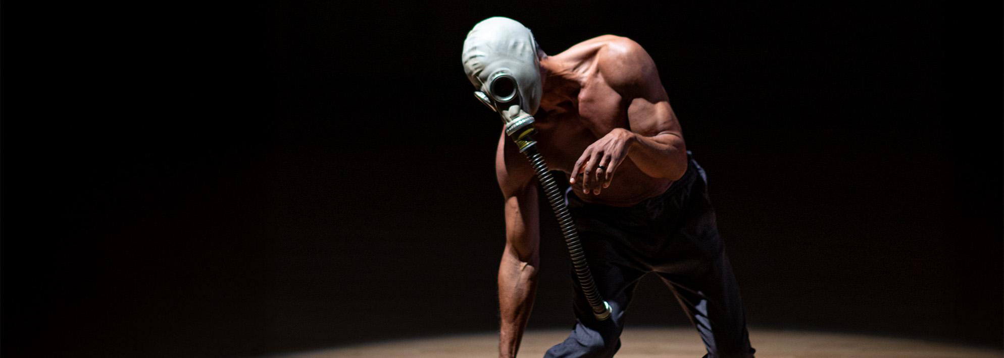 David Rousseve strikes a pose on stage in front of a black backdrop while wearing a gas mask 