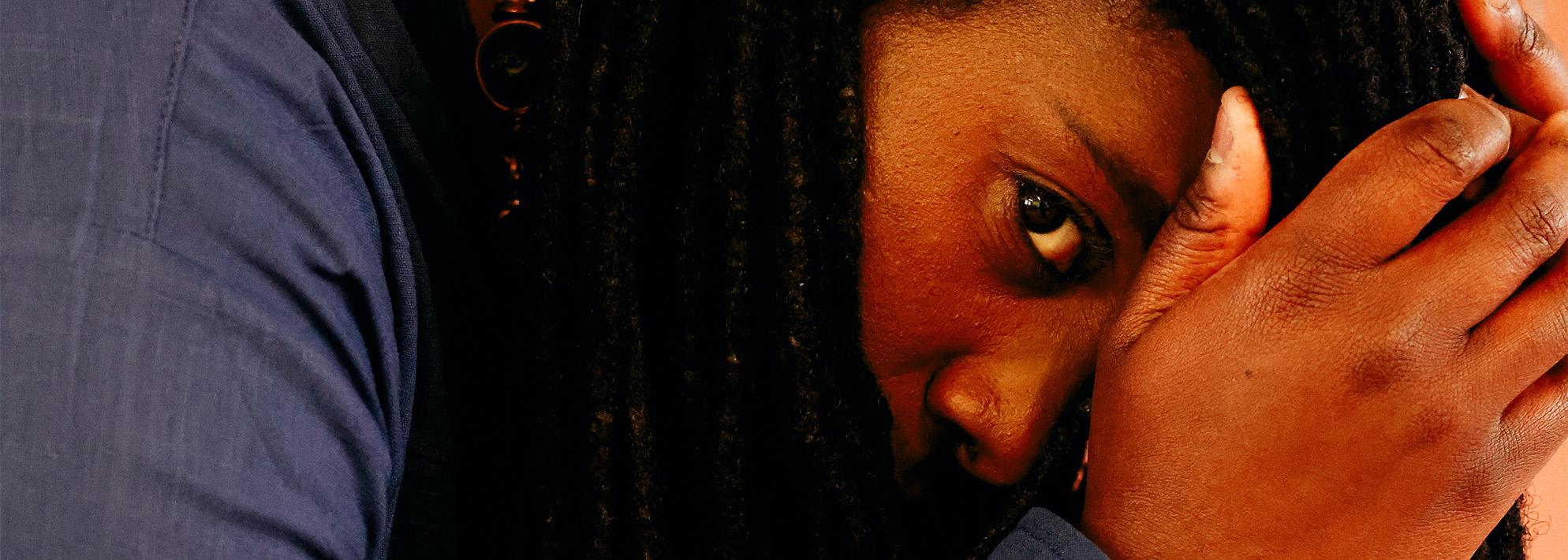 Headshot portrait of Jlin posed in front of a orange background and facing side profile to the camera, covering eyes with her hands