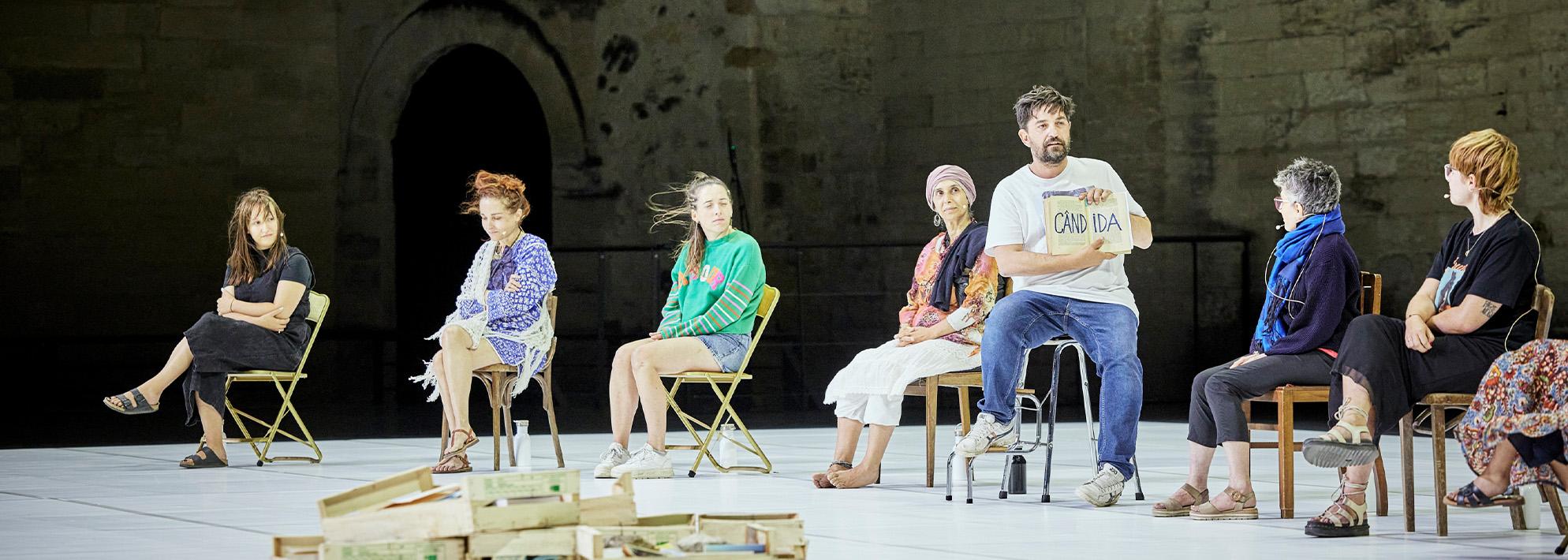 Tiago Rodrigues sits at a chair on stage holding a book with participants seated at separate chairs