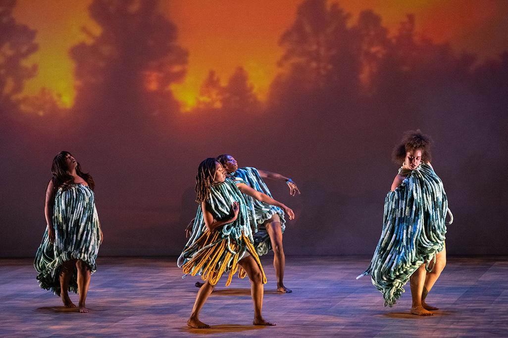 Dancers perform on stage in front of a projection of trees