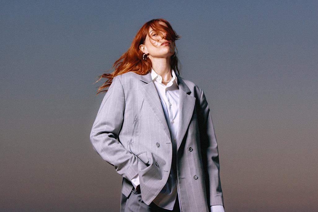 Headshot of Laurel Halo posed in front of a grey background in a grey suit