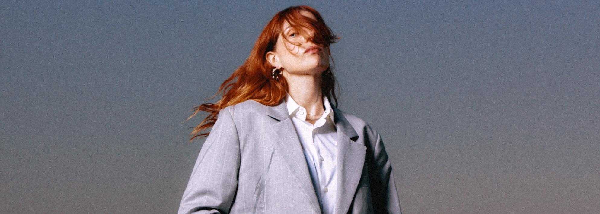 Headshot of Laurel Halo posed in front of a grey background in a grey suit