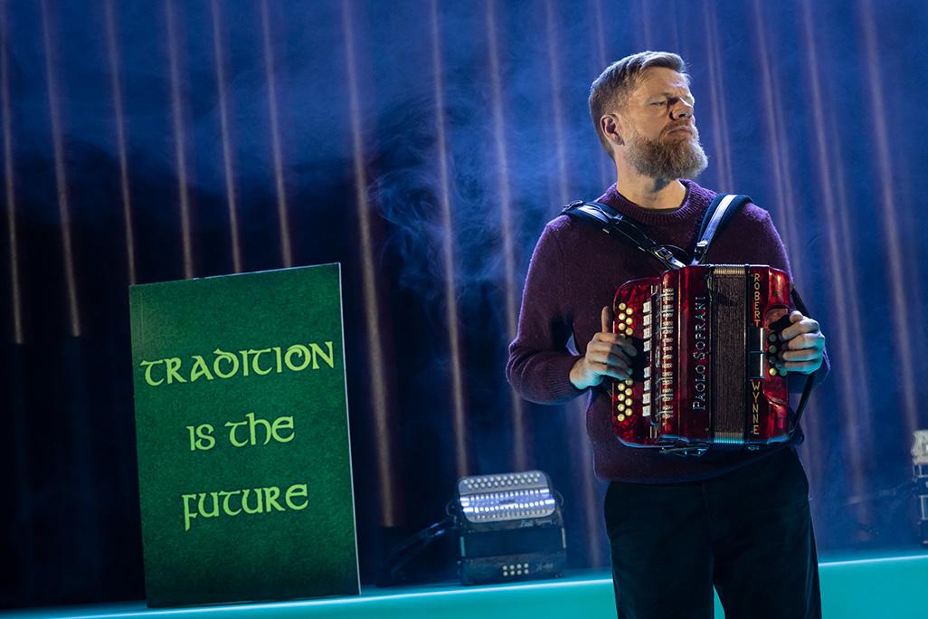 Danny O'Mahony plays the accordion on stage next to a sign reading 'tradition' to the left of him