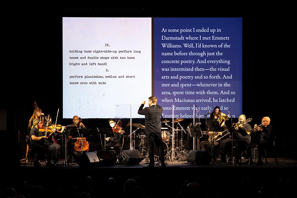 A conductor performs on stage with a 17 piece ensemble and text projection of text and description of music behind them
