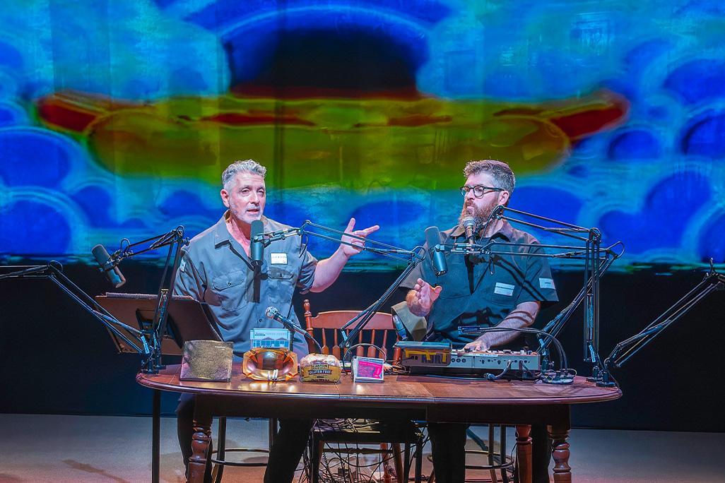 Ain Gordon and Josh Quillen sit at a desk on stage in front of a rainbow background