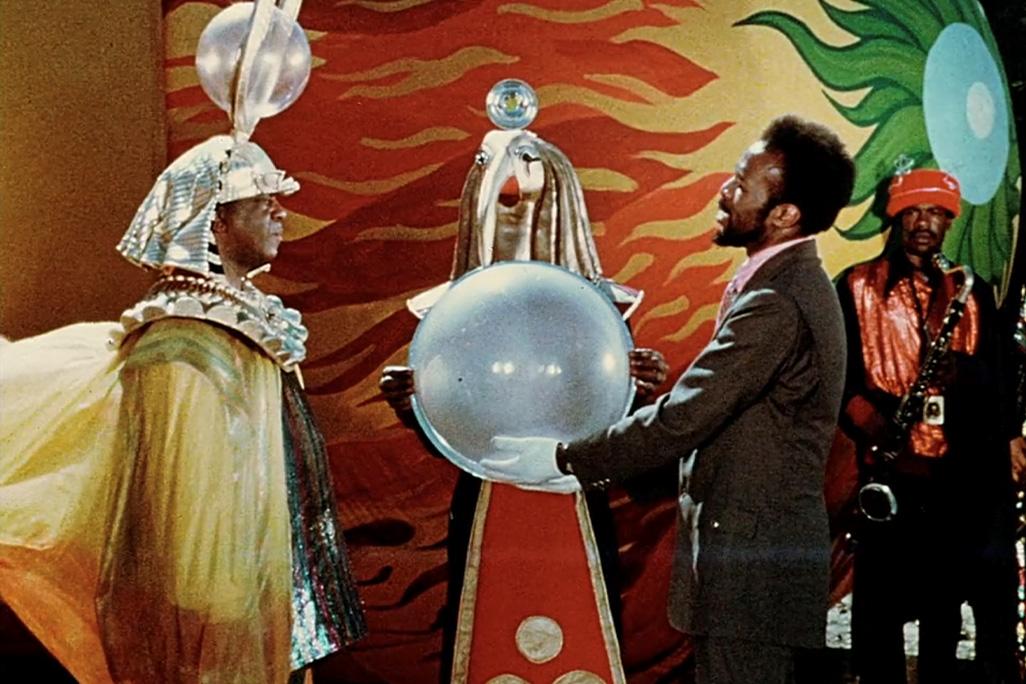 Still from Sun Ra’s ‘Space Is the Place’ featuring two men, one holding a large orb, standing in front of a colorful background