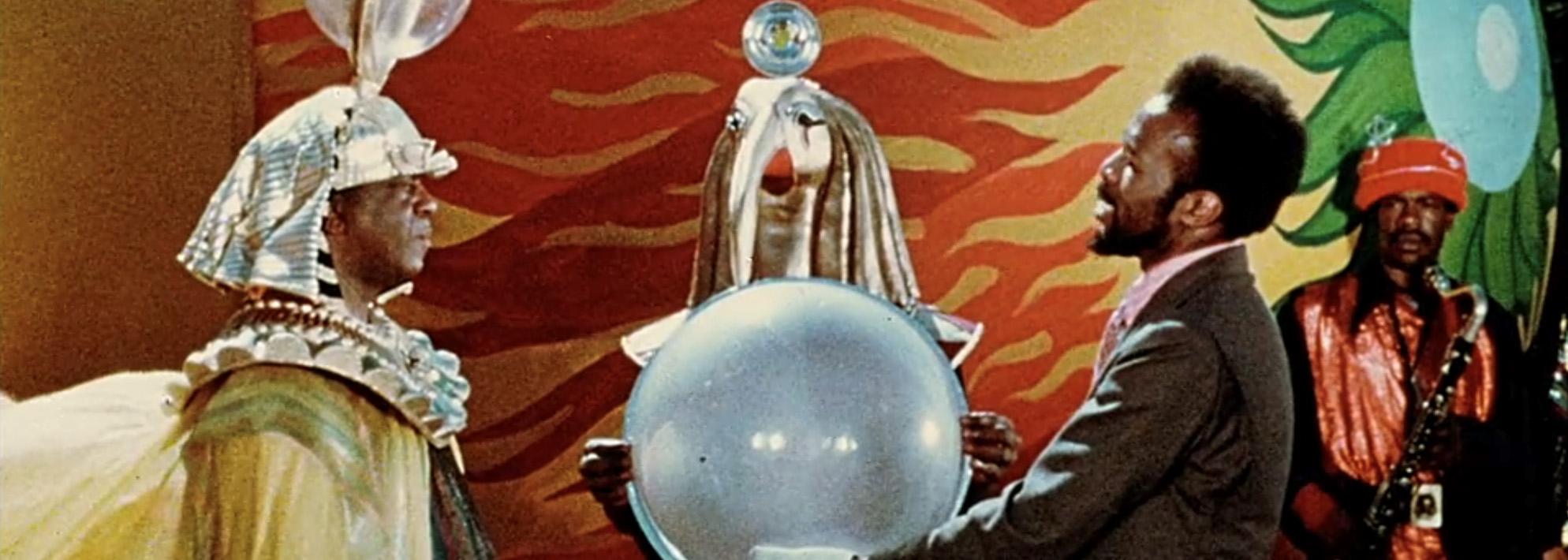 Still from Sun Ra’s ‘Space Is the Place’ featuring two men, one holding a large orb, standing in front of a colorful background