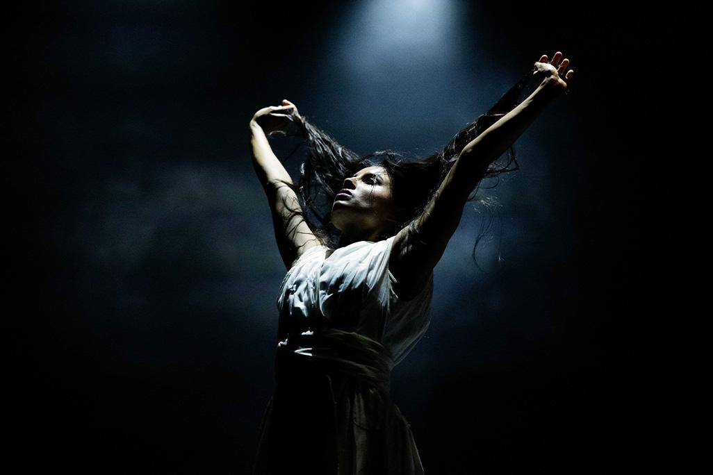 Performer from Akram Khan Company stands on a stage with a dark, smokey backdrop. The performer holds their arms in the air and holds onto their hair.