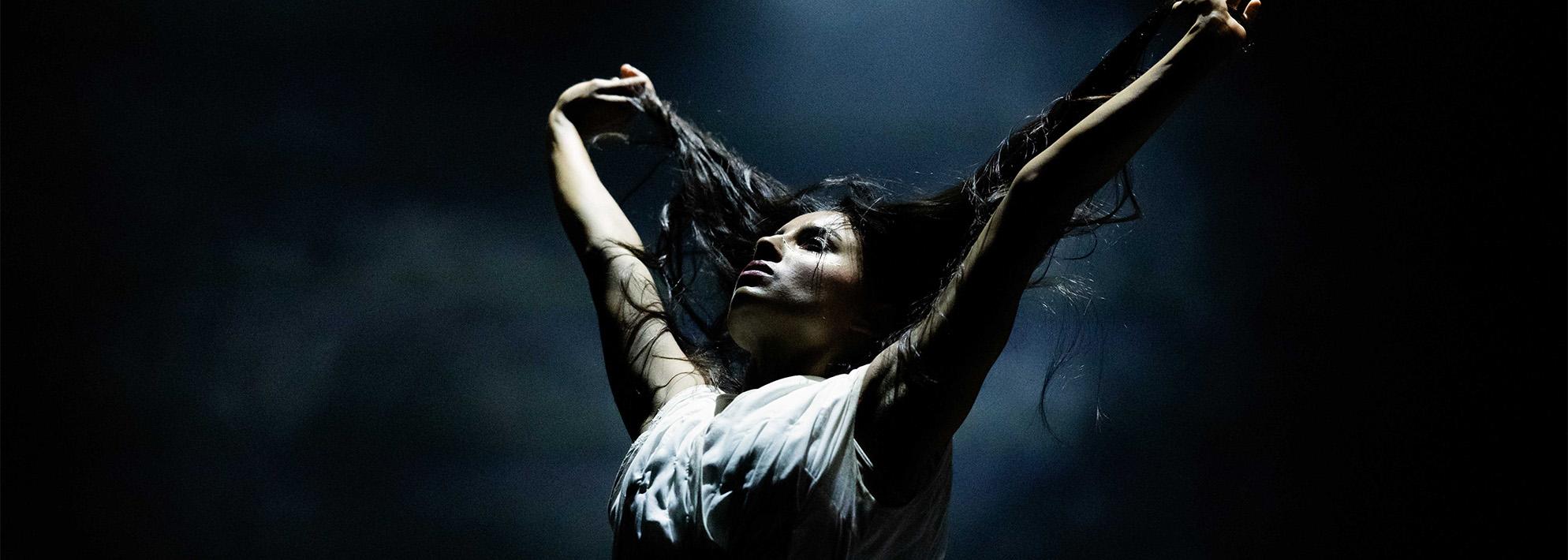 Performer from Akram Khan Company stands on a stage with a dark, smokey backdrop. The performer holds their arms in the air and holds onto their hair.