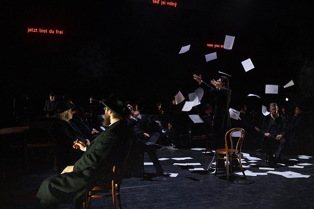 A performer stands on a dark stage wearing a suit while papers fall from the ceiling around them