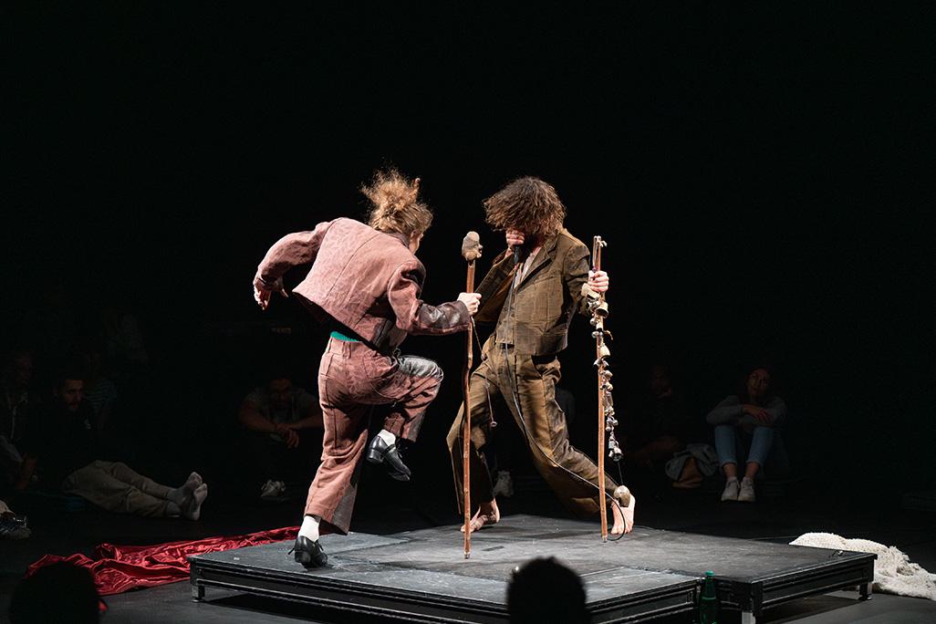 Two performers stand on a black box stage, faced towards each other, mid movement. One dancers holds a standing stick with bells on it.
