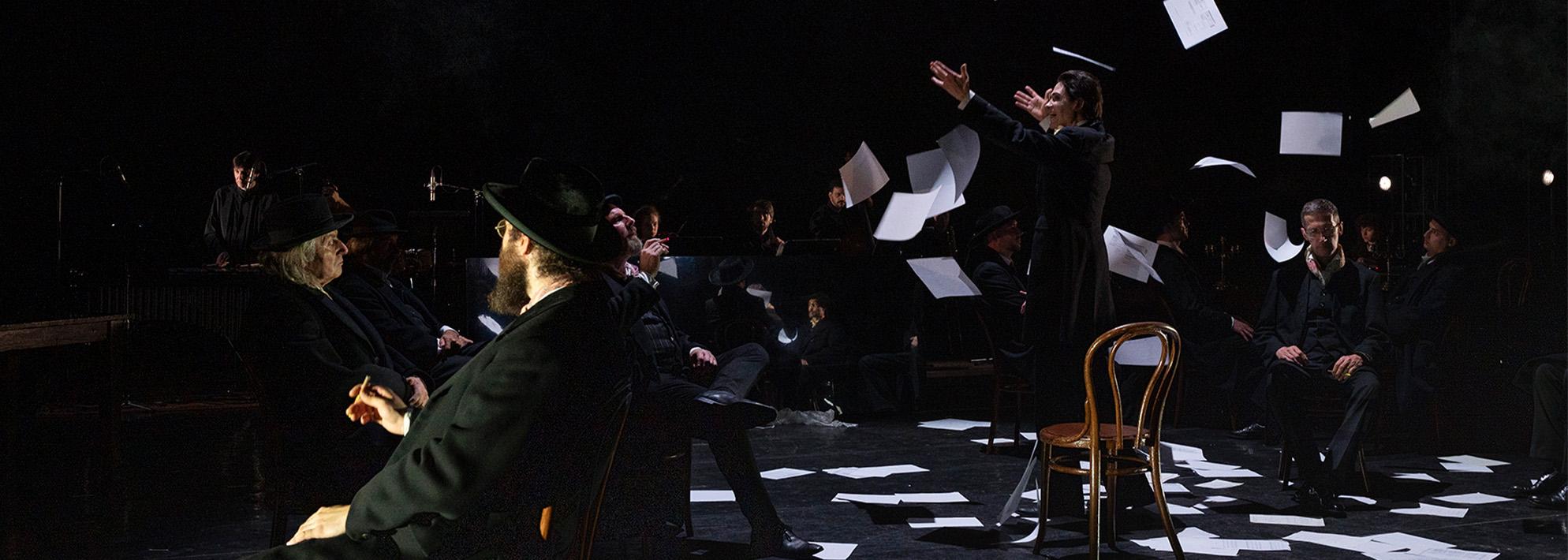 A performer stands on a dark stage wearing a suit while papers fall from the ceiling around them