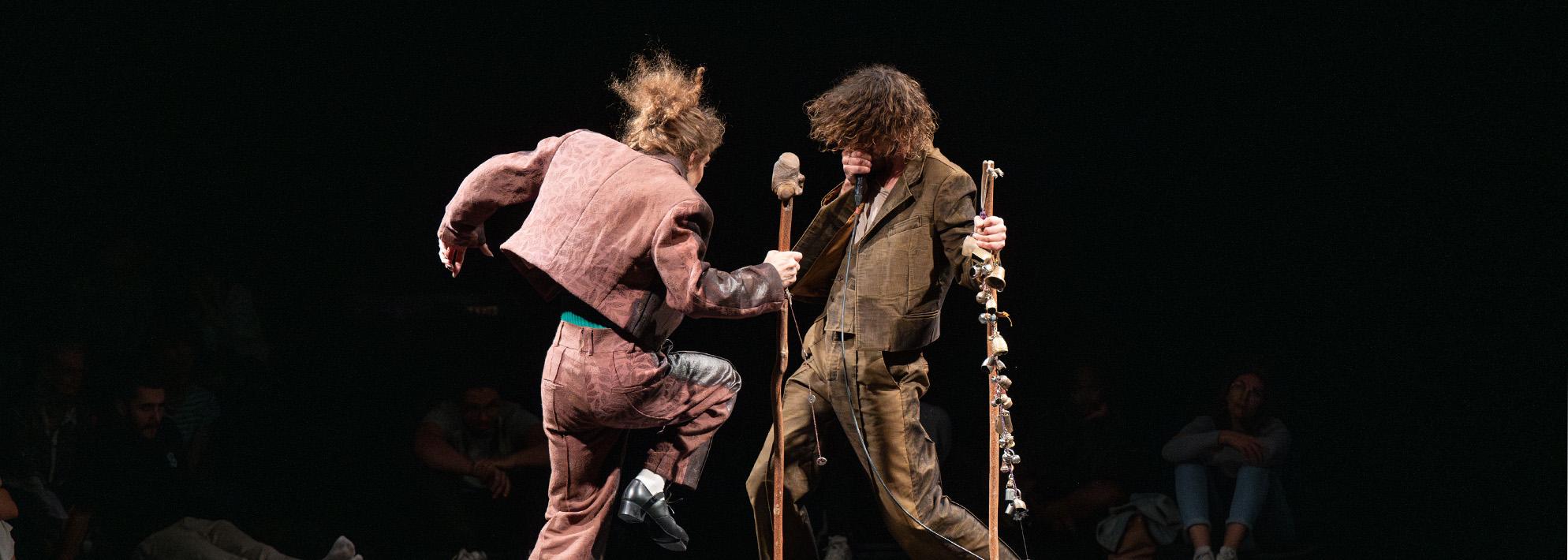Two performers stand on a black box stage, faced towards each other, mid movement. One dancers holds a standing stick with bells on it.